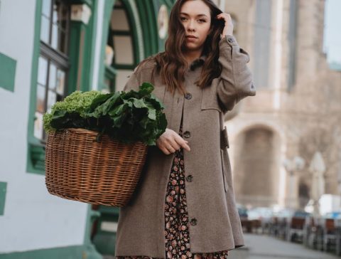 A woman in a brown coat carries a wicker basket of green vegetables while strolling through Erfurt, Germany.